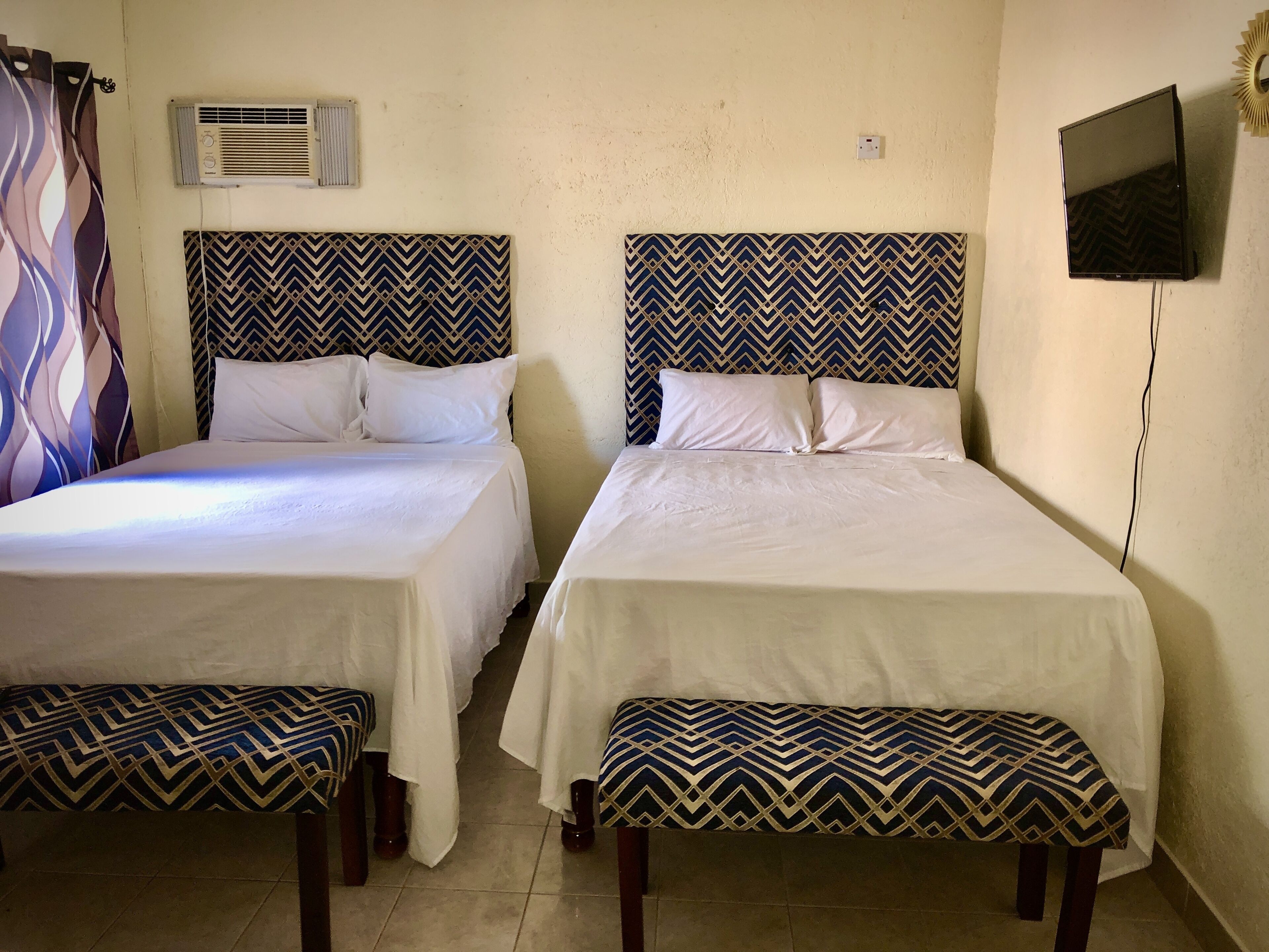 Dos camas individuales con cabeceras y pies de cama iguales, un televisor de pantalla plana montado en la pared y un equipo de aire acondicionado sobre las camas.