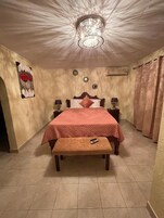 Un dormitorio con cama, mesitas de noche, un banco, una araña y un cuadro en la pared.