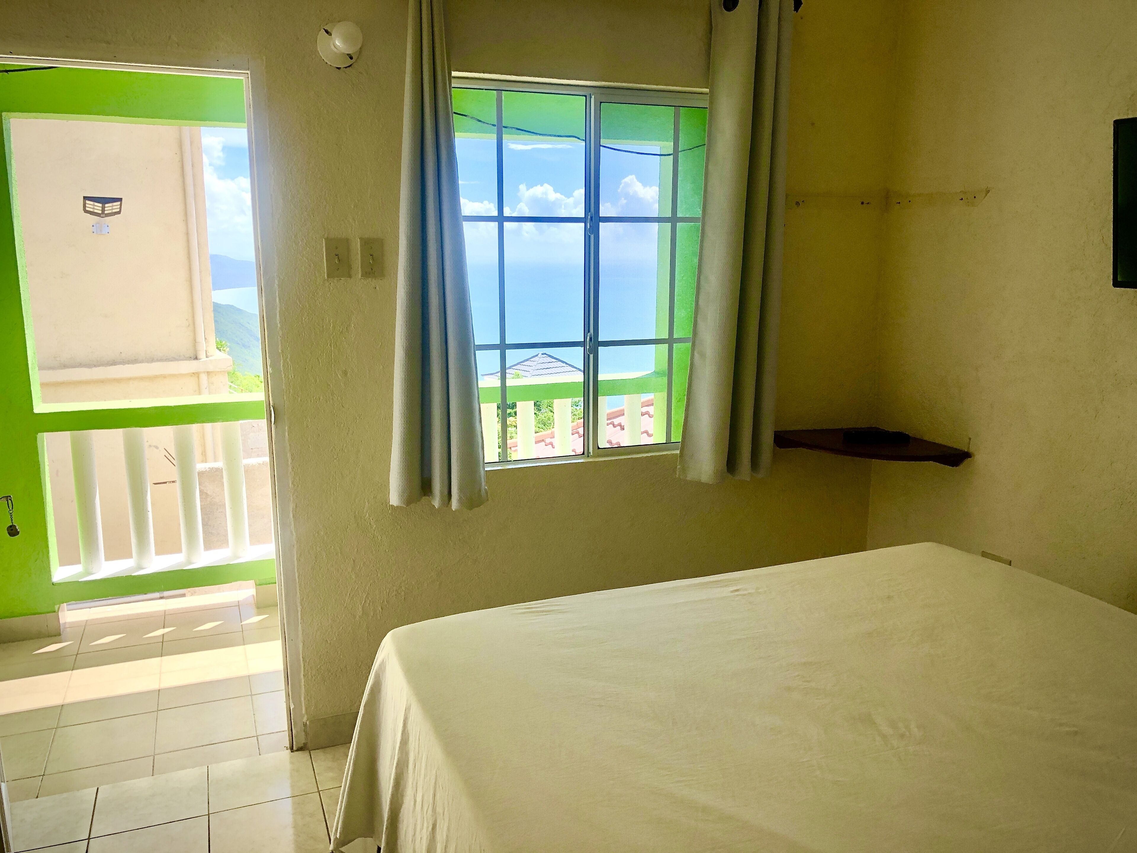 A room with a bed, a window with curtains, a green door, and a view of the outside.