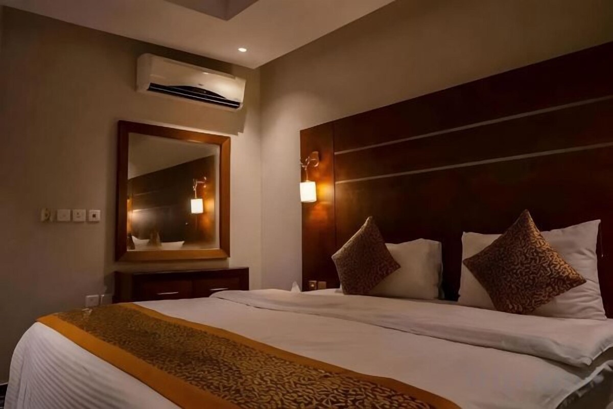 A hotel room with a large bed, a headboard with lights, a mirror, and an air conditioning unit.