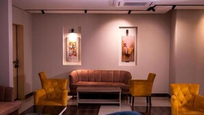 A modern living room with a brown sofa, two yellow armchairs, a blue armchair, and a glass coffee table. There are two wall-mounted artworks and a ceiling light fixture. - Ahlin 2 Serviced Apartment (Riyadh)