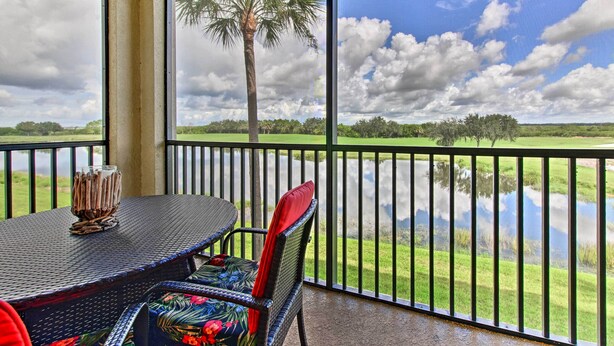 A balcony with a table and chairs, overlooking a body of water and a grassy landscape with a palm tree. - Screened Patio & Resort Perks: Bradenton Condo! (Bradenton)