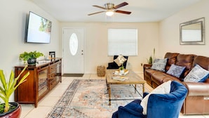 Un salon avec un canapé en cuir marron, un fauteuil bleu, une table basse en bois, un ventilateur de plafond, un téléviseur fixé au mur et un tapis à motifs.