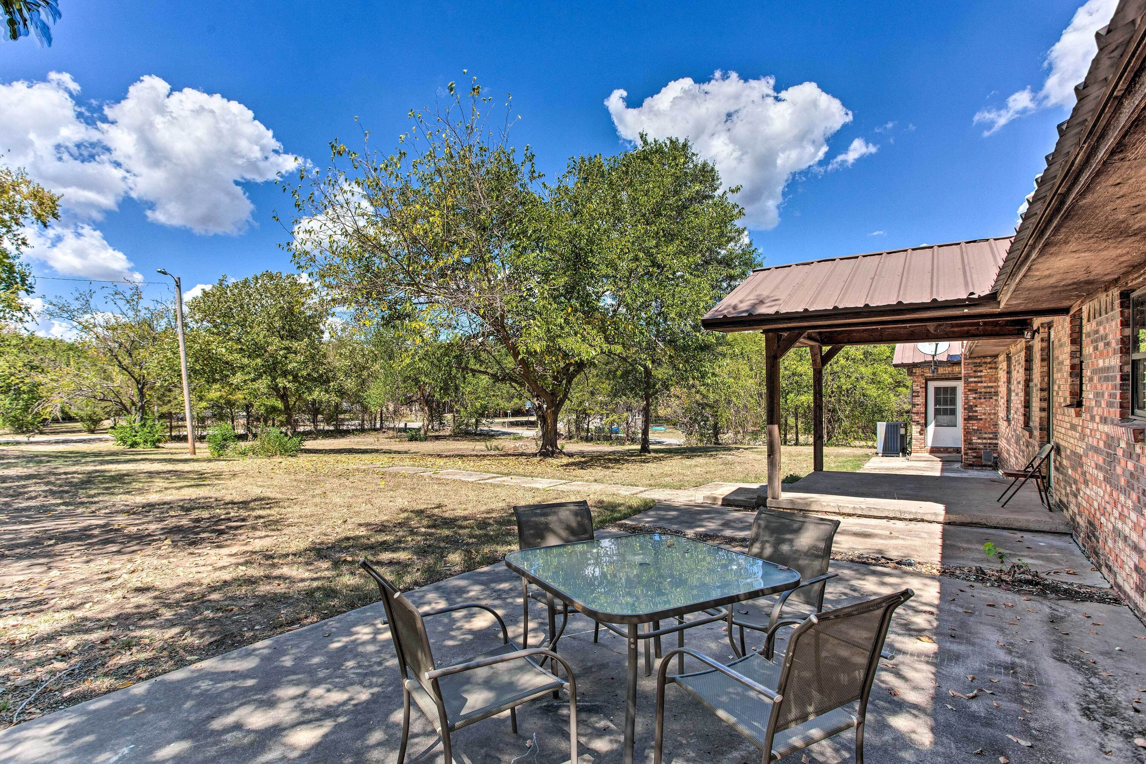 A patio with a glass table and chairs, a brick building, and trees in the background.