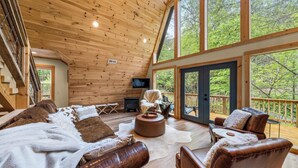 Ein geräumiges Blockhauskaminzimmer mit einer großen Holzbalkendecke, einer gemütlichen Sitzecke mit Ledersofas, Kamin und Fernseher.