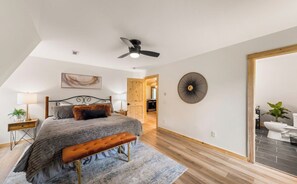 A bedroom with a bed, a bench, a ceiling fan, a wall art piece, and a door leading to another room.
