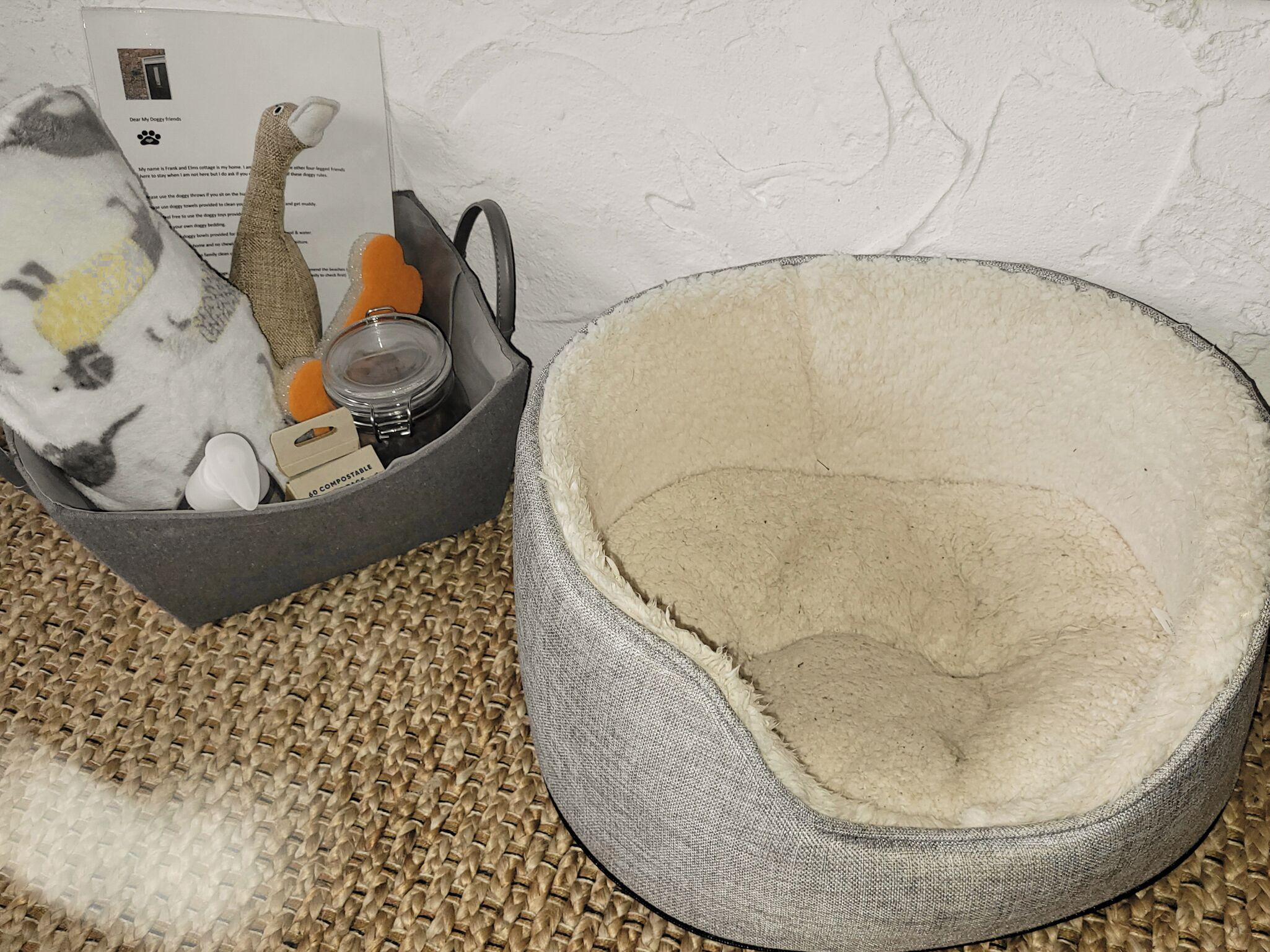 A grey pet bed with a fleece interior, a woven mat, and a grey basket containing a cat toy, a small container, and a tag.