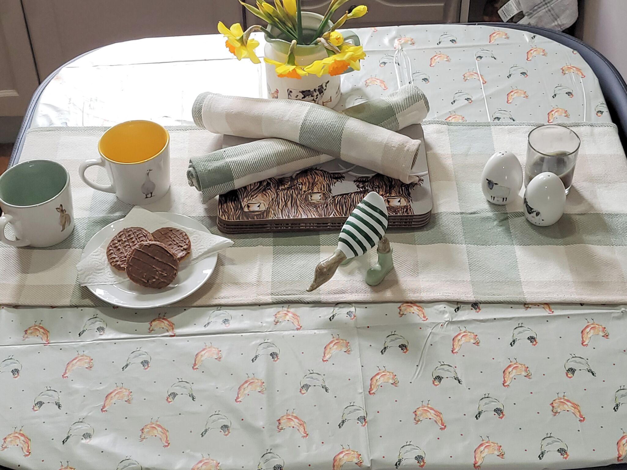 Une table bien rangée, recouverte d’une nappe à carreaux, avec une assiette de biscuits, une tasse, un vase de fleurs, un jouet rayé et un plateau décoré.