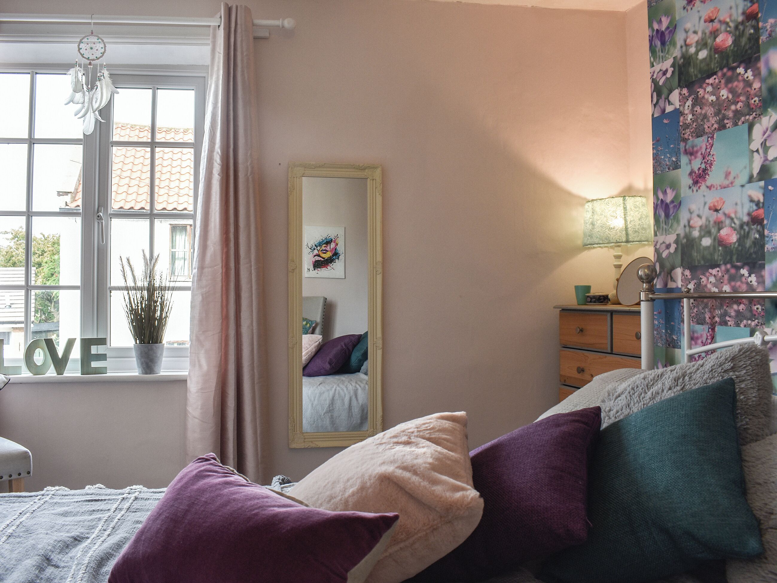 A bedroom with a bed, pillows, a dresser, a mirror, a window with curtains, a plant, and a wall with floral wallpaper.