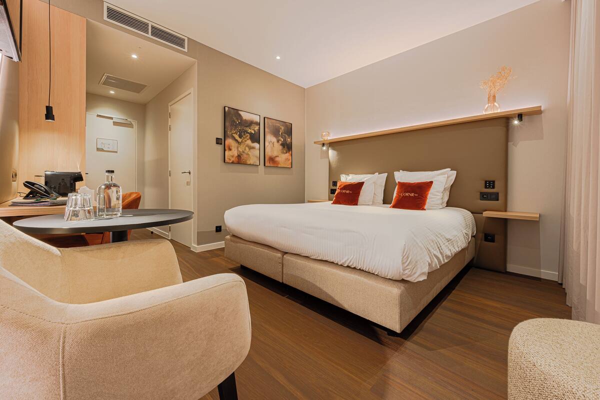 A modern hotel room with a bed, a desk, and a chair. There are two paintings on the wall and a vase on the nightstand.