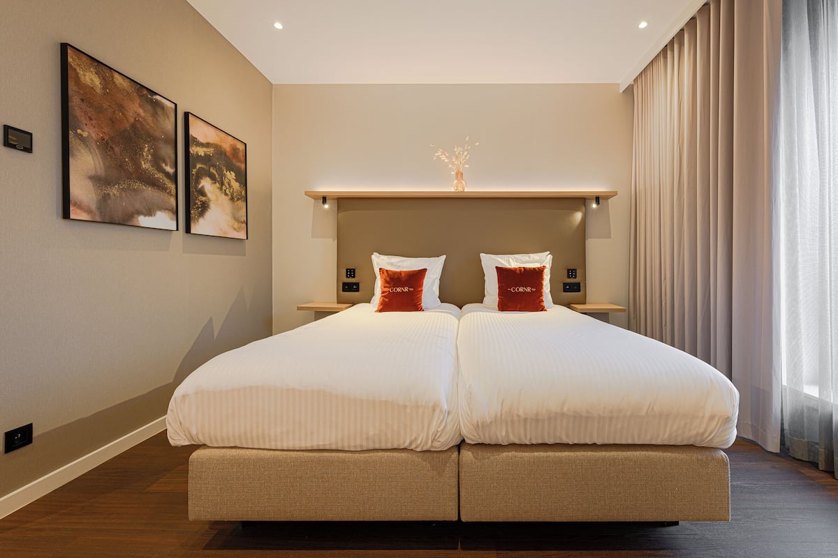 A double bed with white linens and two red pillows, a bedside table with a vase, and two abstract paintings on the wall.