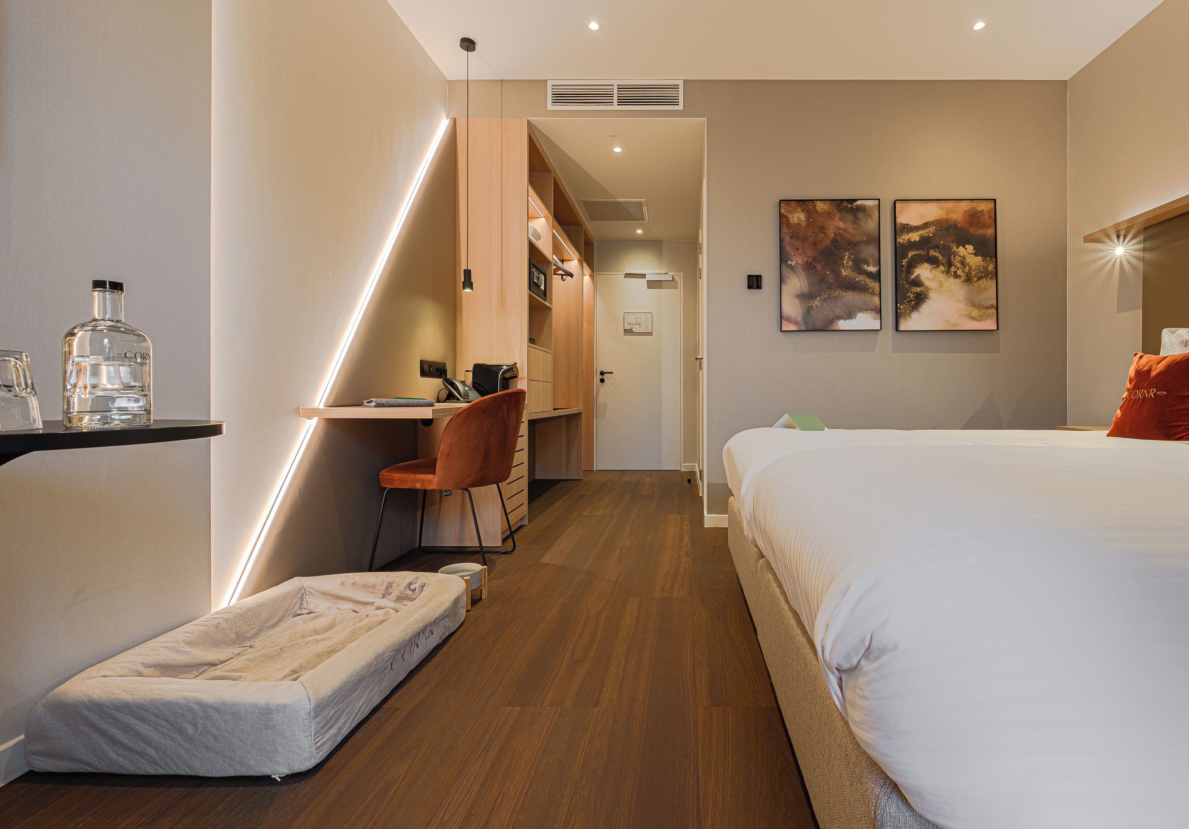 A modern hotel room with a bed, a desk, and a chair. There is a small bedside table with a bottle and glasses. The room has wooden flooring and a white wall.