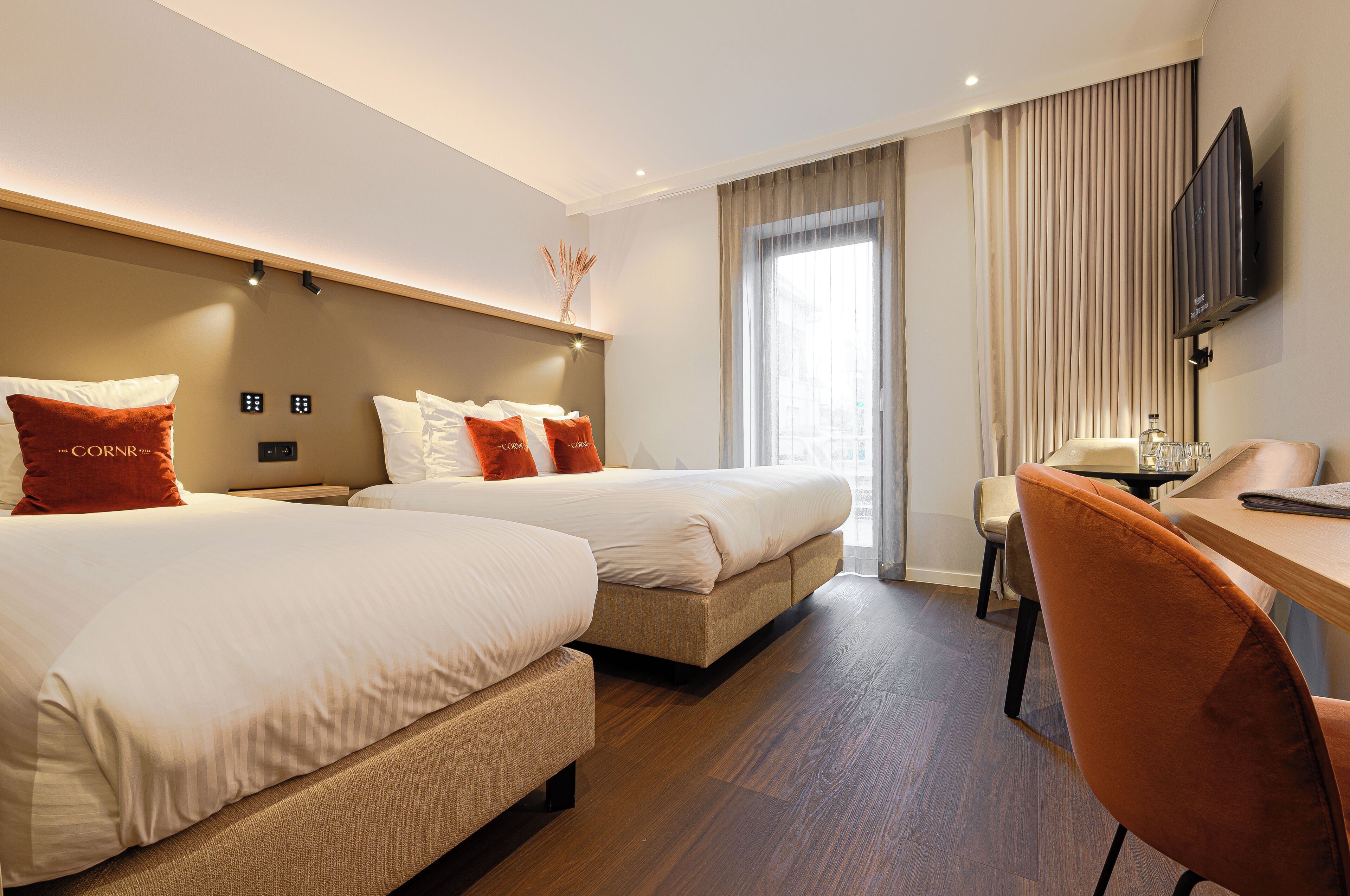 A hotel room with two beds, a desk, a chair, a TV, and a window with curtains.