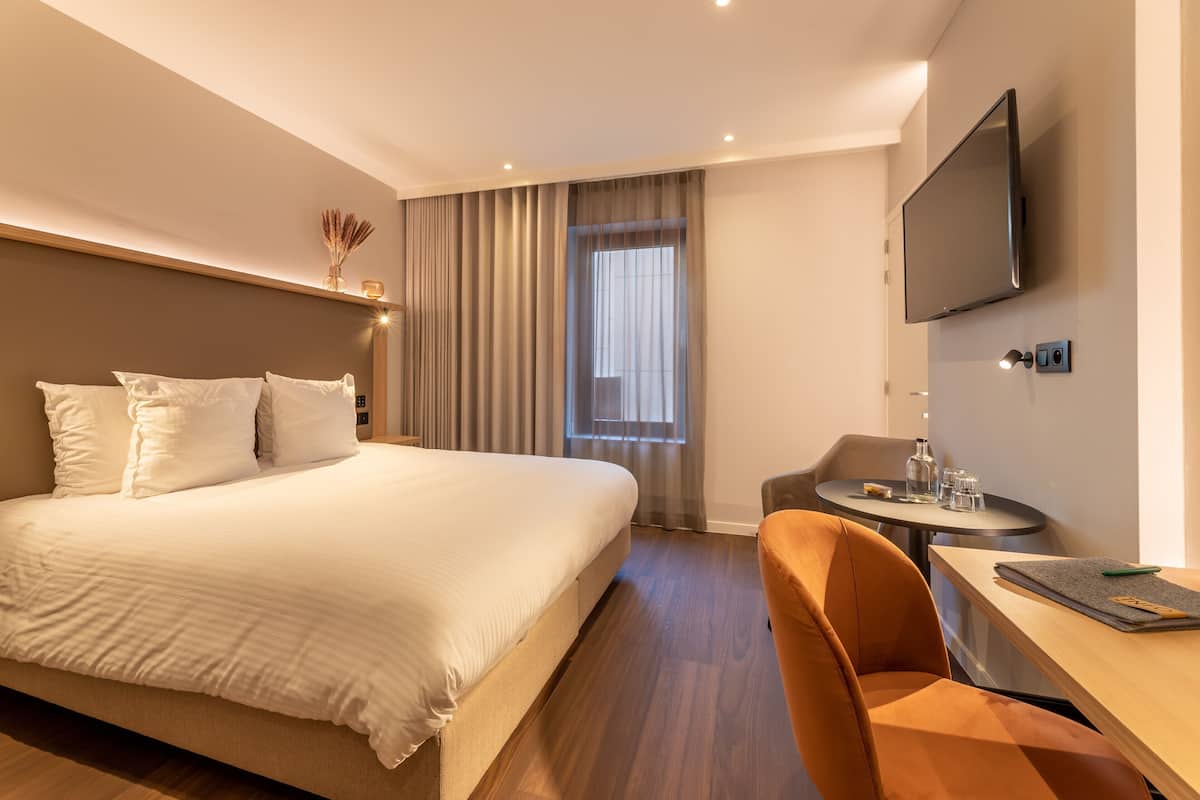 A hotel room with a bed, a desk with a chair, a TV, and a window with curtains.