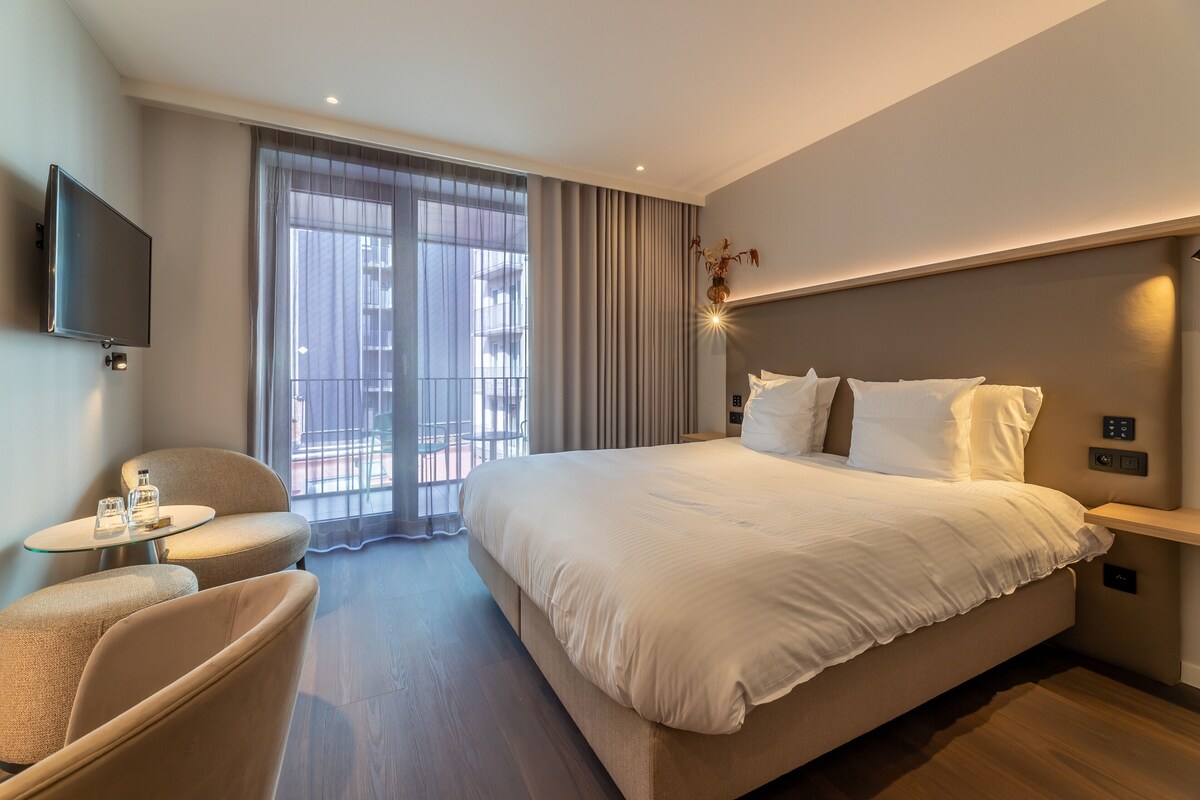 Ein modernes Hotelzimmer mit einem großen Bett, einem an der Wand befestigten Fernseher, einem kleinen runden Tisch und einer Sitzecke mit einem Sessel.