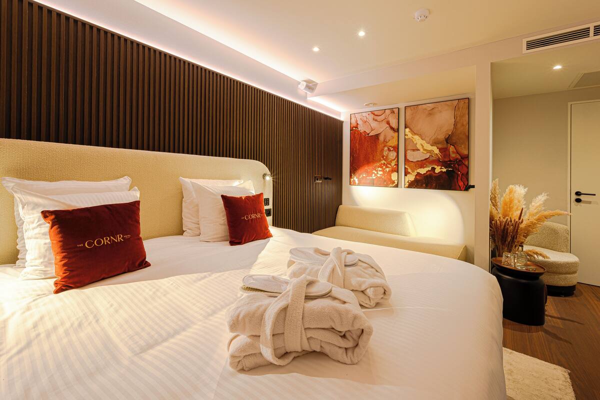 A hotel room with a large bed, two red pillows, a white headboard, and a painting on the wall.