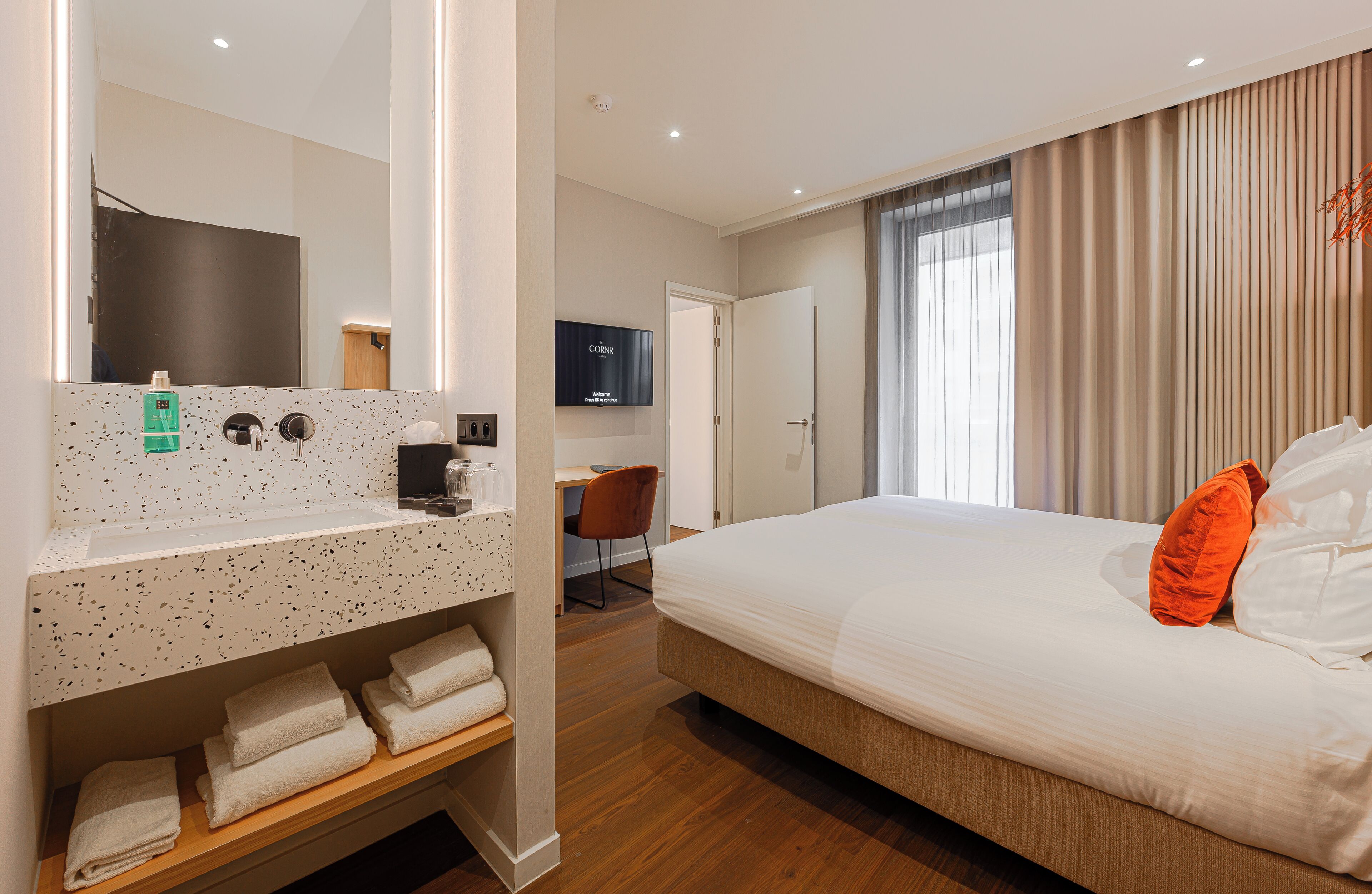 Habitación de hotel con una cama grande, un escritorio de madera, un televisor y un baño con espejo y perchero.