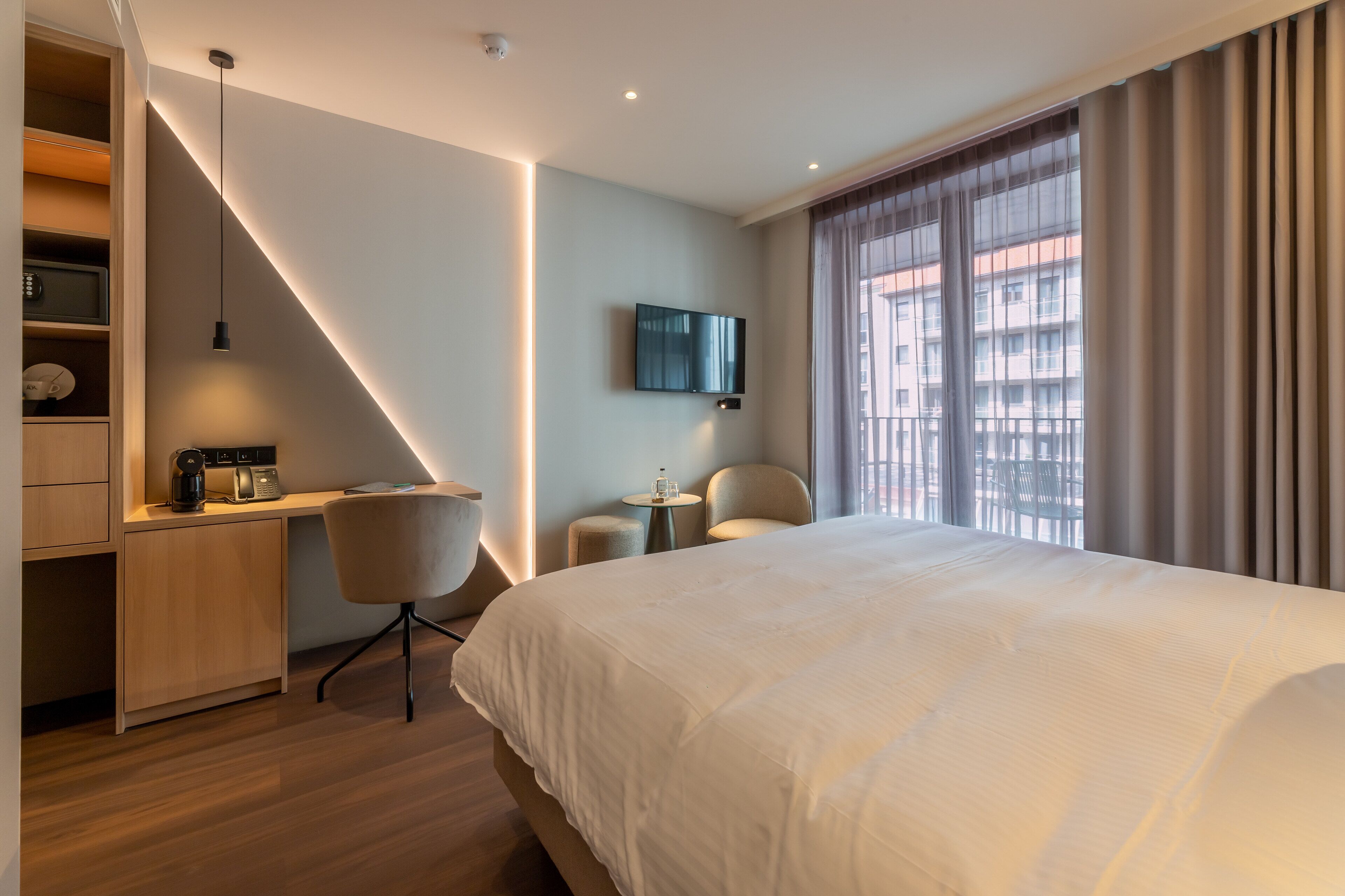 A modern hotel room with a large bed, a desk with a chair, a television, and a view of the city through large windows.