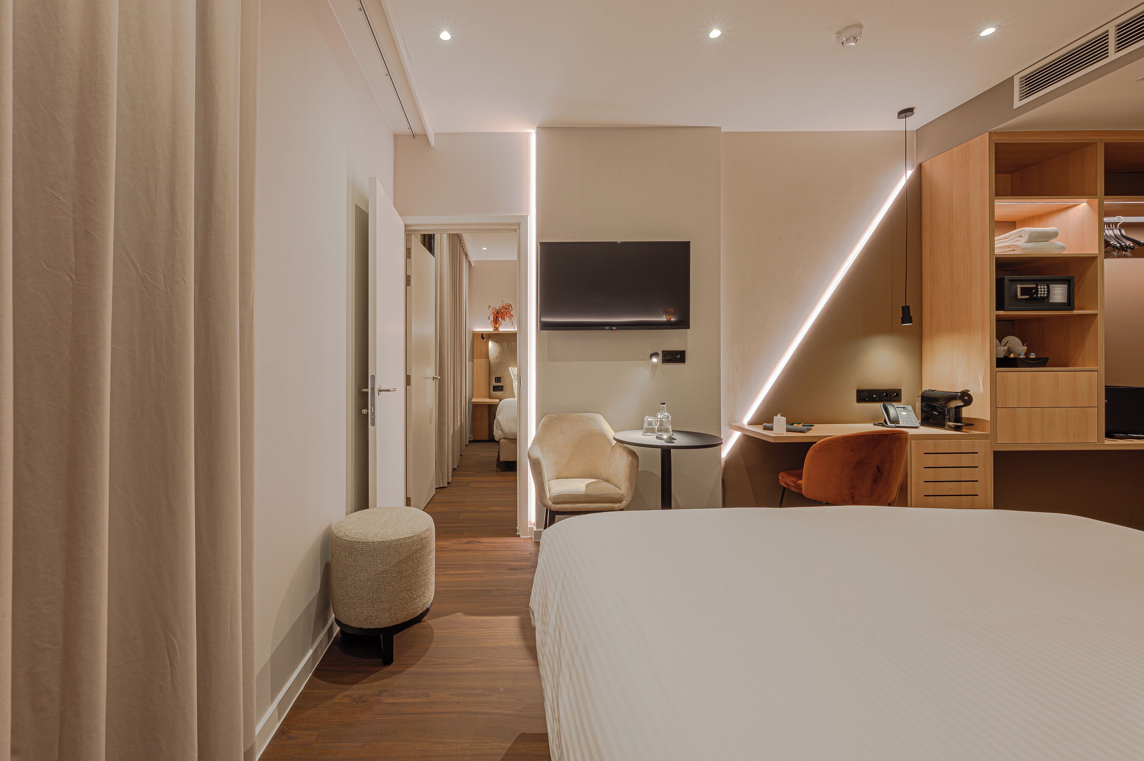 Ein modernes Hotelzimmer mit einem großen Bett, einem Schreibtisch mit Computer, einem Sessel und einem kleinen Hocker.