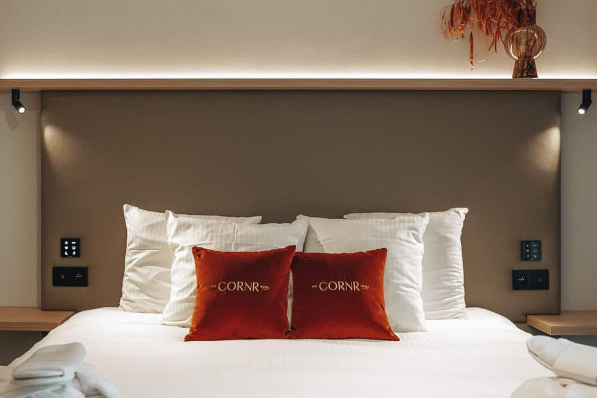 A neatly made bed with white linens and two red pillows labeled "The CORNR HOTEL".