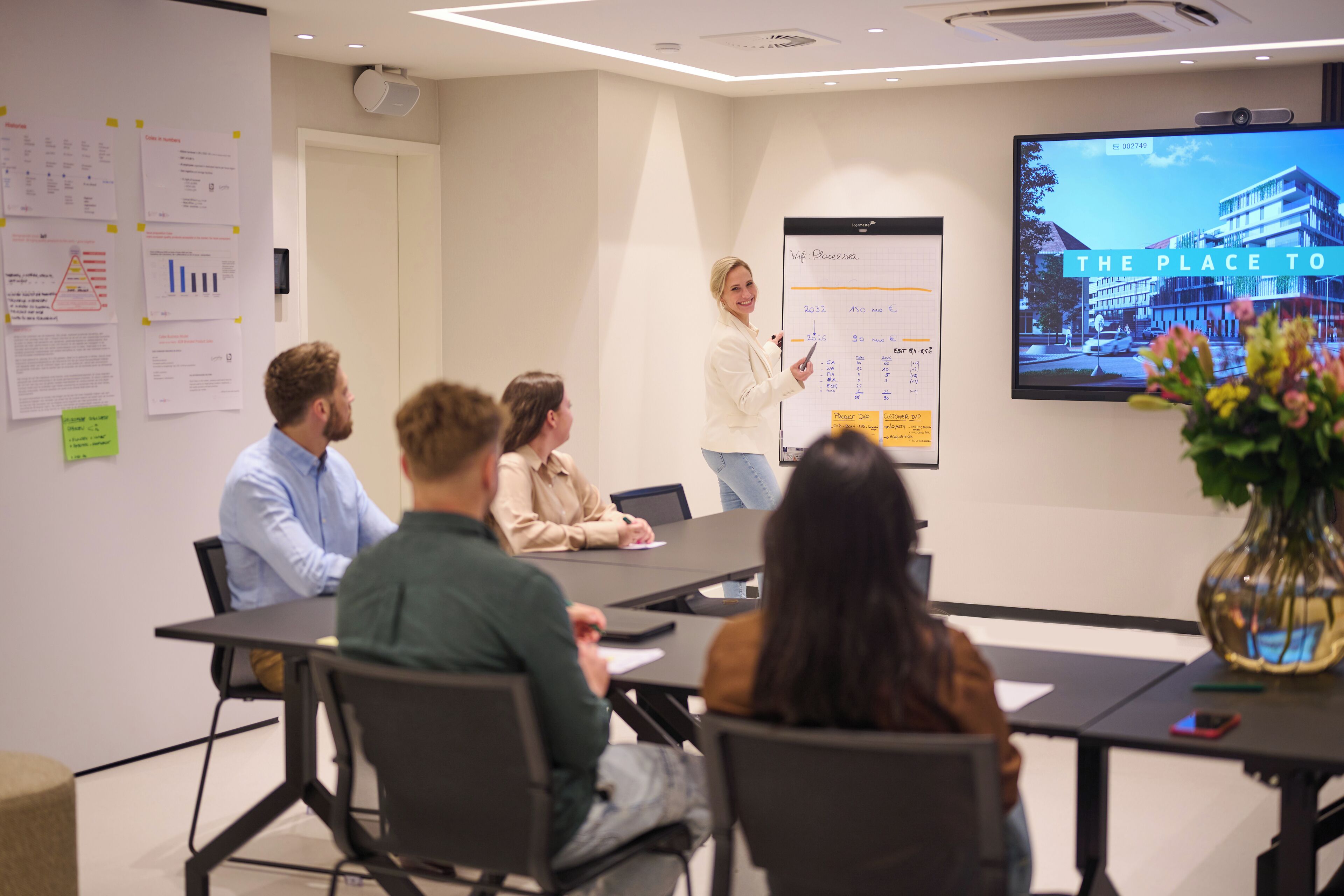 A conference room with a presentation screen displaying a building image and a whiteboard with text and charts.
