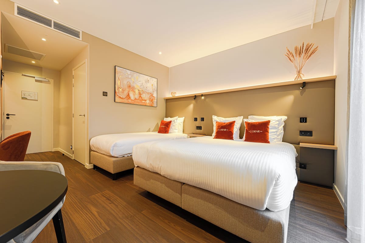 A hotel room with two beds, a small table, and a chair. There is a painting on the wall and a decorative vase on the nightstand.
