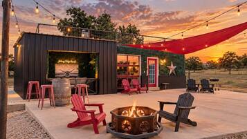 An outdoor seating area with red chairs, a fire pit, and string lights against a sunset backdrop.