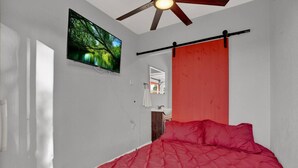 Ein Schlafzimmer mit Bett, Deckenventilator, Fernseher, einer roten Tür und einem Fenster.