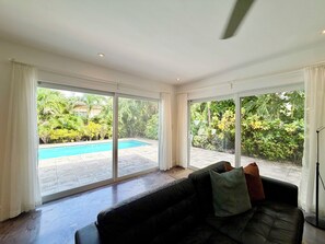 A spacious living room with a black leather sofa, large sliding glass doors leading to a pool and garden.