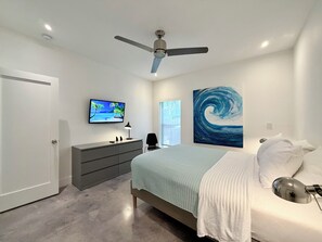 Un dormitorio con cama, televisor, cuadro, ventilador de techo y ventana.
