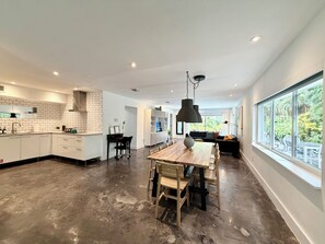 A modern kitchen with white cabinetry, a dining area with a wooden table, and a large window offering a view of greenery. - Heated pool-2 streets from beach-downtown-4 bedroom-3bath (Fort Lauderdale)
