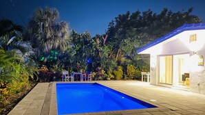 Une piscine dotée d’une terrasse carrelée, entourée d’une végétation luxuriante et bordée d ’un bâtiment blanc avec une porte en verre.