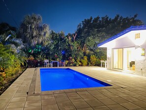 Una piscina con borde de baldosas, rodeada de exuberante vegetación y un edificio blanco con puerta de cristal.