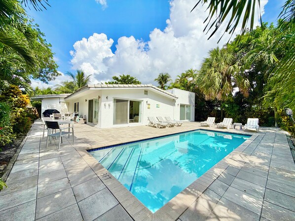 A swimming pool with lounge chairs, a covered patio area, and a building with a tiled roof.