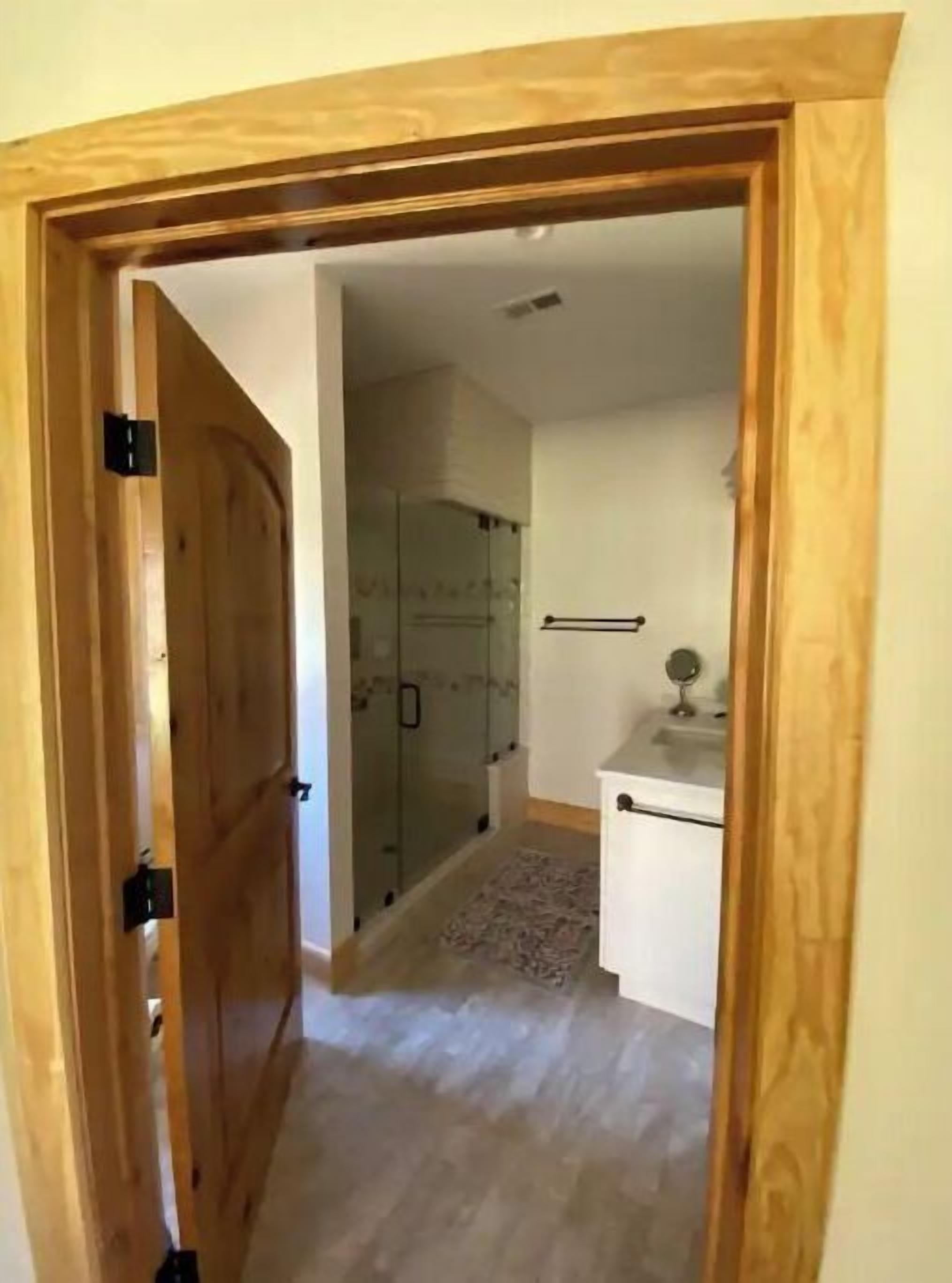 Puerta de madera que da a un baño con cabina de ducha de vidrio y un lavamanos pequeño.