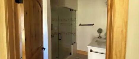 Puerta de madera que da a un baño con cabina de ducha de vidrio y un lavamanos pequeño.