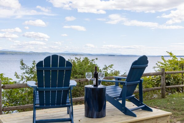 Deux fauteuils Adirondack sont placés face à un lac, sur une terrasse en bois, et une bouteille de vin est posée sur une table.