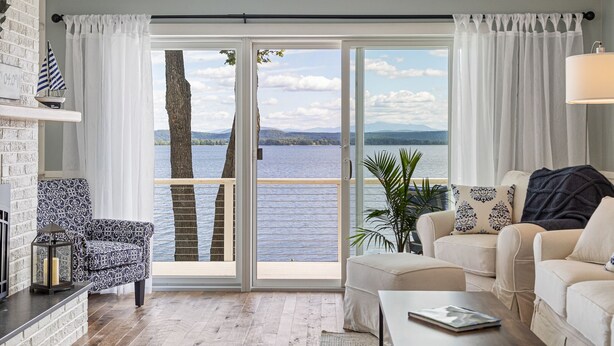 A living room with a large sliding glass door leading to a lake view, a fireplace, a patterned armchair, a sofa, and a coffee table. - Tranquil lake house in VT w/ breathtaking views! (Grand Isle)