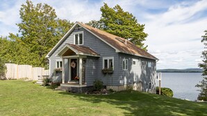 A single-story house with a sloped roof, a front porch with a welcome sign, and a view of a lake. - Tranquil lake house in VT w/ breathtaking views! (Grand Isle)