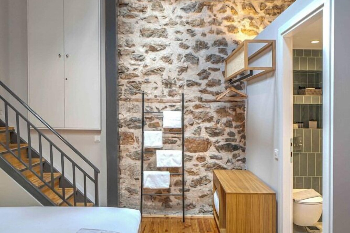 Un dormitorio con suelo de madera, pared de piedra, cama, armario, banco y baño visible a través de una puerta abierta.