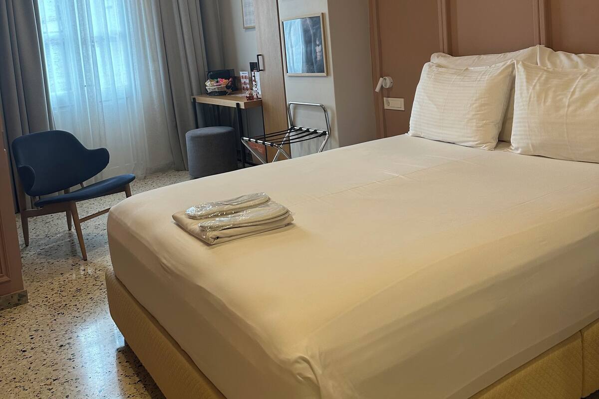 Habitación de hotel con una cama grande, un escritorio con silla, una ventana con cortinas y una estantería con cuadros enmarcados.