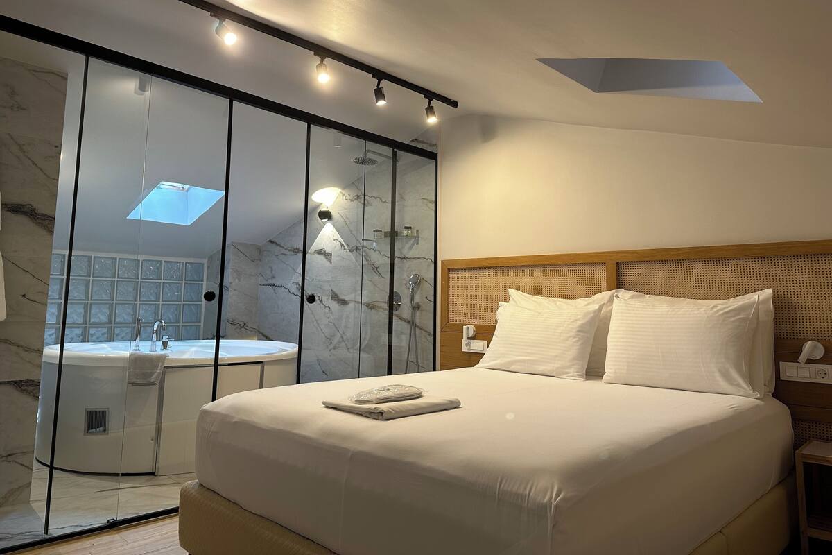 Una habitación de hotel moderna con una cama grande, un cabecero de madera, una cabina de ducha de vidrio y un tragaluz.