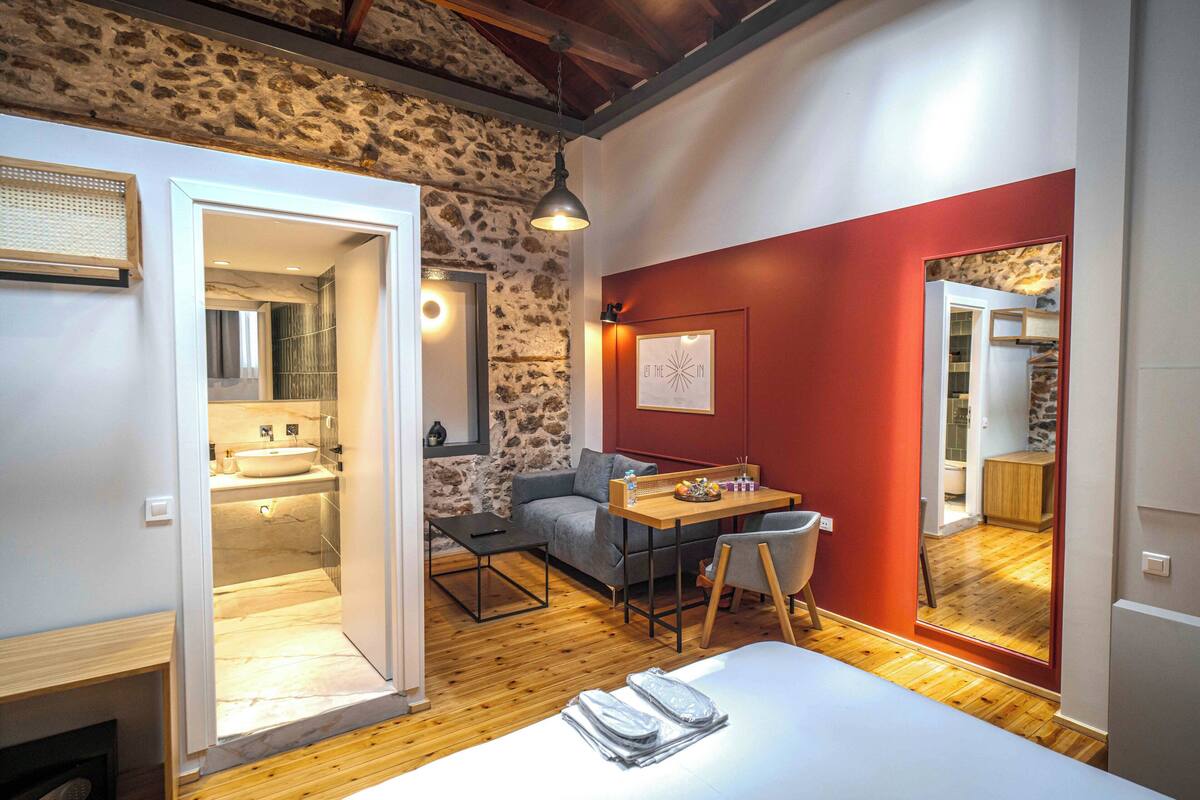 Un dormitorio moderno con suelo de madera, una cama, un escritorio y una silla. Se ve un baño a través de una puerta abierta.