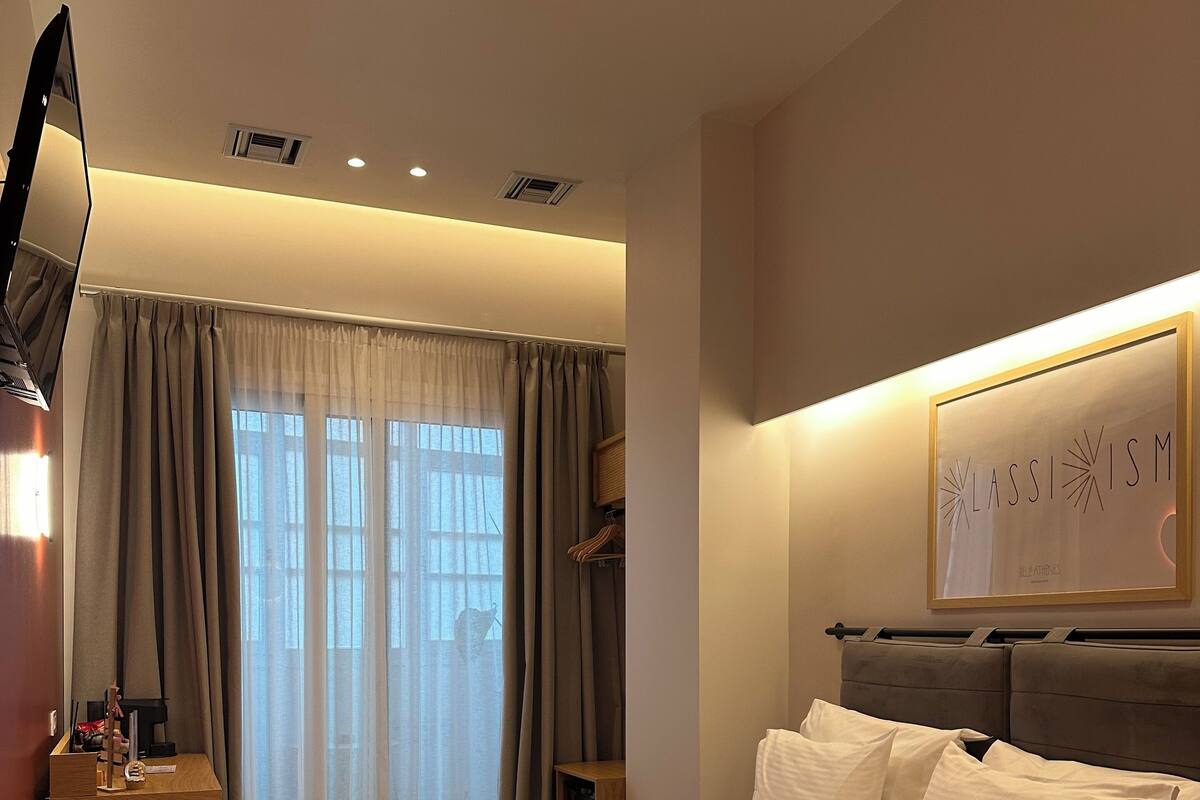 Una habitación de hotel con una cama grande, un televisor de pantalla plana montado en la pared, una mesita de noche con lámpara y una ventana con cortinas.