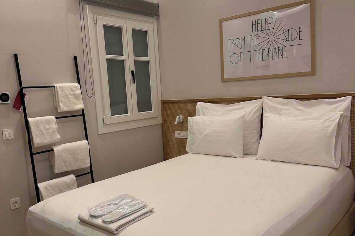 Una cama bien tendida con sábanas blancas, un perchero con toallas dobladas, una cita enmarcada en la pared y una ventana con vidrio satinado.