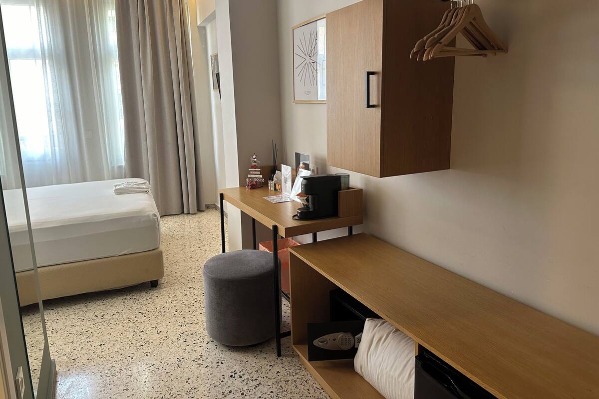 Habitación de hotel con cama, escritorio, silla, pequeño reposapiés, armario y perchero.