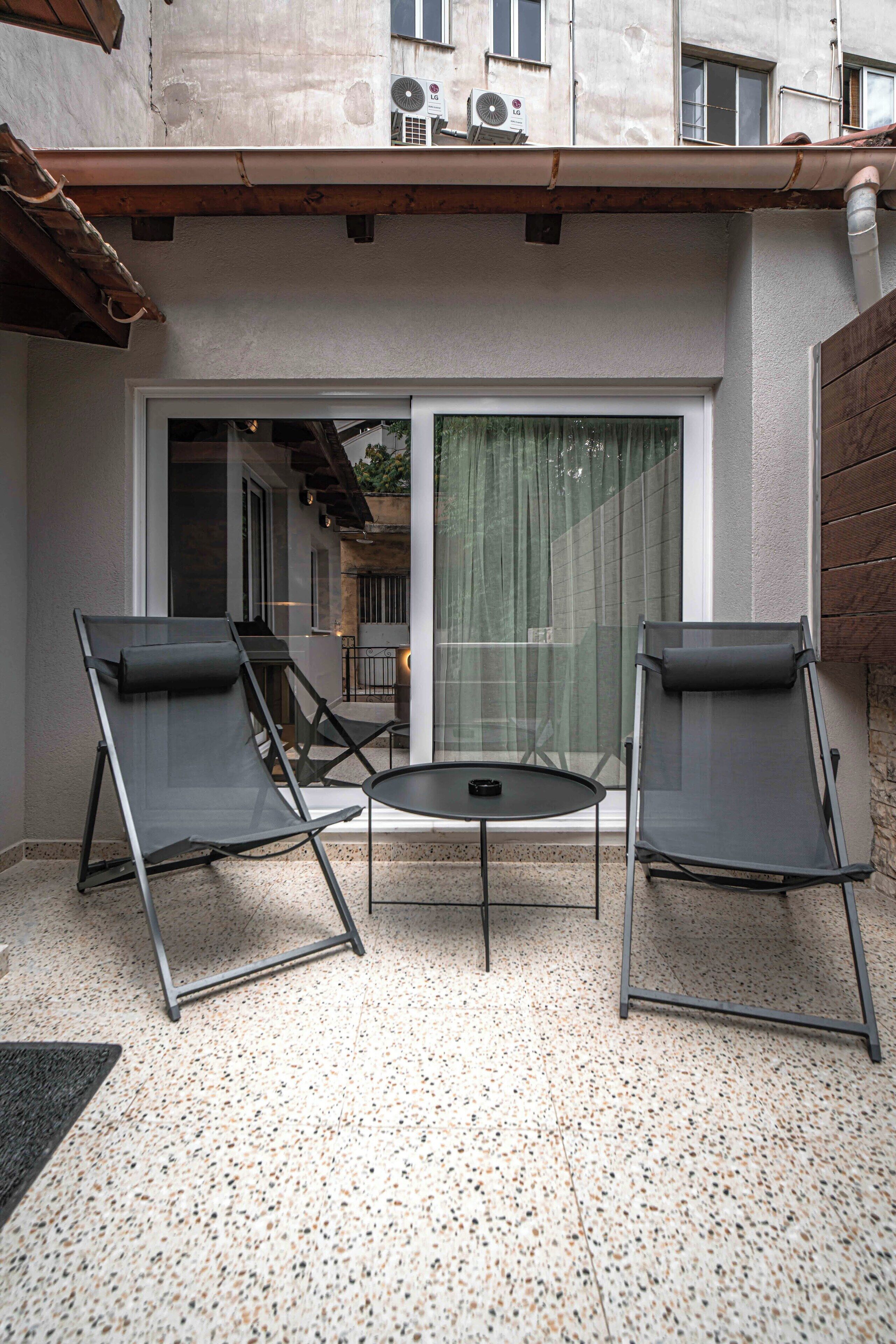 An outdoor patio with two lounge chairs and a small table, a sliding glass door, and a building with a tiled roof and an air conditioning unit.