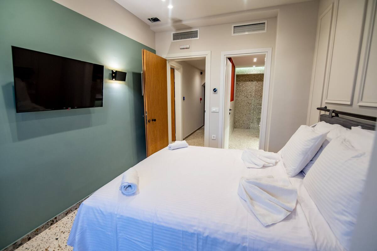 Habitación de hotel con cama, televisión montada en la pared, armario de madera y baño visible a través de una puerta abierta.