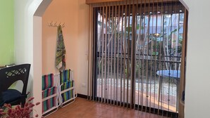 Ein Zimmer mit Fliesenboden, einem schwarzen Stuhl, einem Tisch mit einer Vase getrockneter Blumen, einer Bank mit gestreiften Kissen und Blick durch ein vergittertes Fenster auf einen Außenbereich mit Pflanzen und einem Tisch.