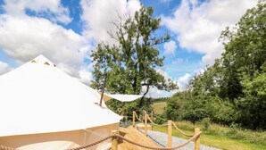 Ein weißes Zelt mit Holzterrasse und Seilbegrenzung, umgeben von Bäumen und einem klaren Himmel.